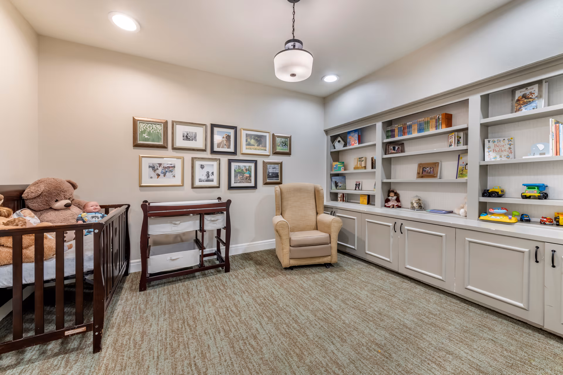 A cozy room with a crib containing large teddy bears and a baby doll, a changing table with storage bins, a beige armchair, and built-in white shelves filled with books, toys, and decorative items. The walls are adorned with framed pictures, and the room is well-lit with ceiling lights.