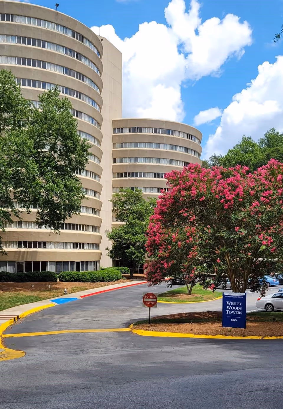 Exterior view of Wesley Woods Towers, showing two curved multi-story buildings surrounded by trees and landscaping. A driveway with a 'Do Not Enter' sign and a blue sign with the facility name is visible in the foreground under a partly cloudy sky.