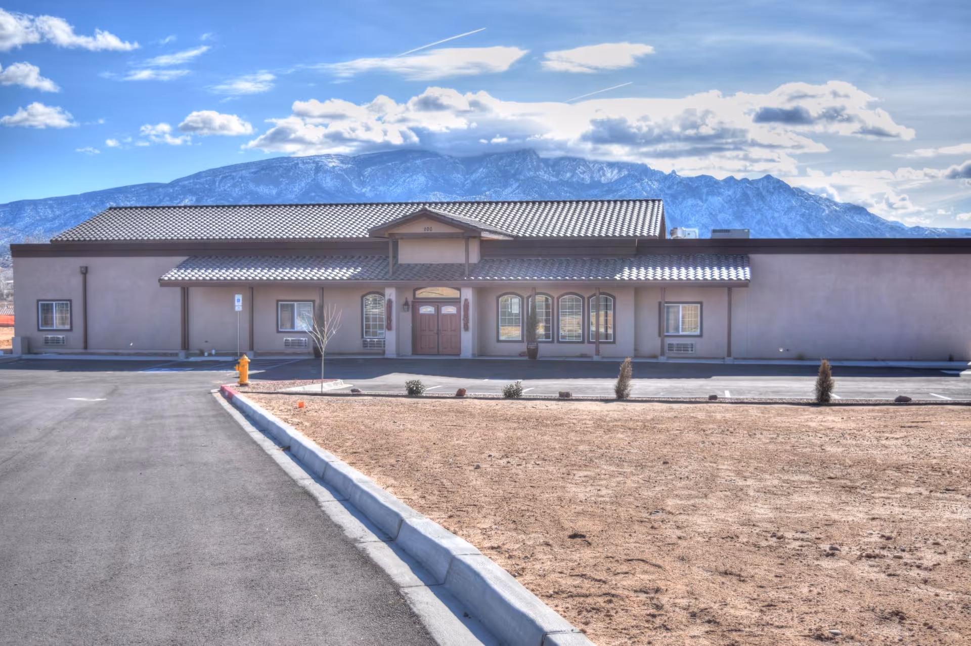 Front exterior view of a single-story building with a tiled roof and multiple windows, set against a backdrop of snow-capped mountains under a partly cloudy sky. The building has a paved driveway and a small landscaped area with sparse vegetation in front.