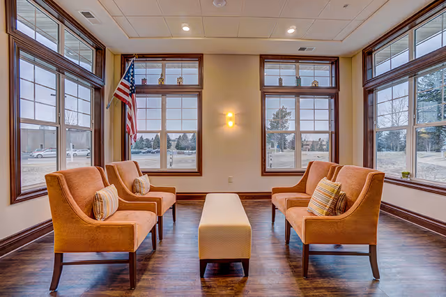 A bright sitting area with four orange upholstered armchairs arranged in two pairs facing each other, separated by a long beige ottoman. The room has large windows with wooden frames on three sides, allowing natural light to fill the space. An American flag is displayed near one window, and a wall sconce provides additional lighting. Outside, trees and parked cars are visible.