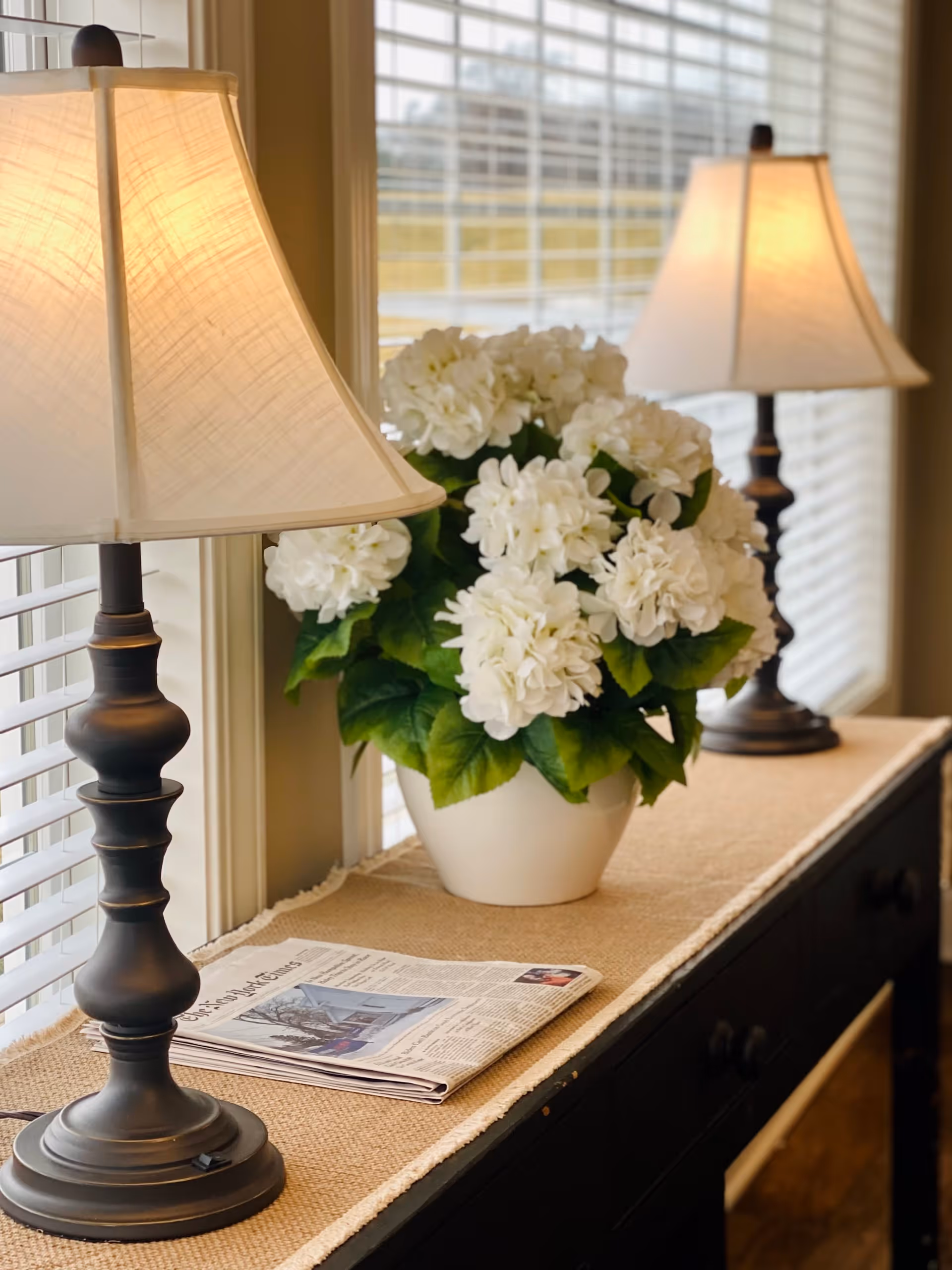 A black console table with a beige runner, holding a white vase filled with white flowers and two lit table lamps with beige lampshades, positioned in front of windows with white blinds. A folded newspaper is also placed on the table.