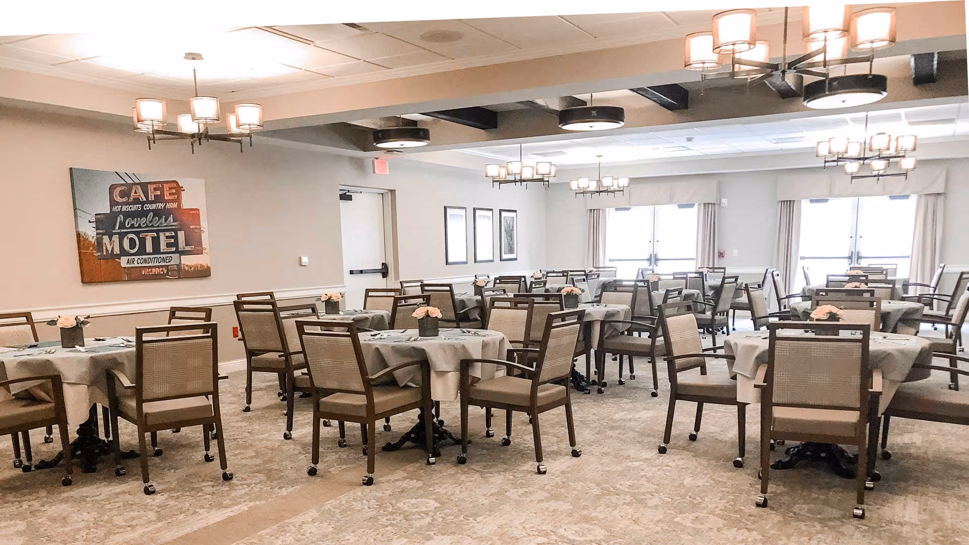 A spacious dining room with multiple tables covered with beige tablecloths, each surrounded by four wooden chairs with cushioned seats. The room is well-lit with modern ceiling lights and has large windows with curtains allowing natural light to enter. A vintage-style wall art piece is visible on the left wall.