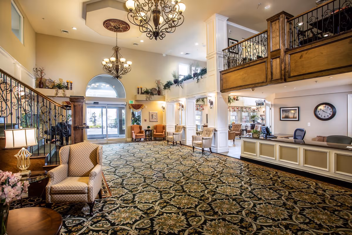 Spacious and elegant senior living facility lobby with patterned carpet, multiple armchairs, a reception desk, decorative chandeliers, and a staircase with ornate railing. Large windows and glass doors allow natural light to brighten the area.