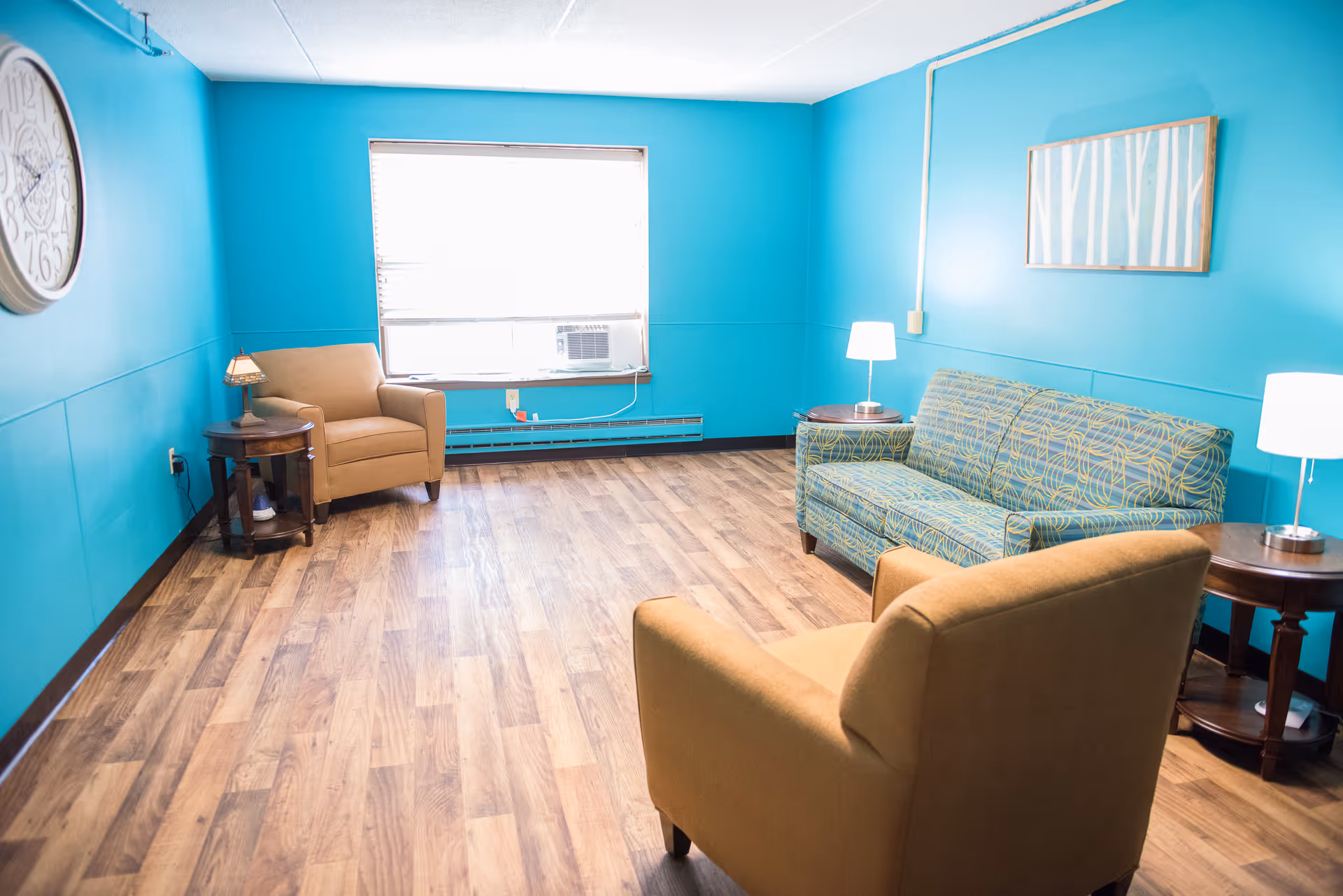 Bright turquoise-walled lounge with wood flooring, two armchairs, a patterned sofa, side tables and lamps.