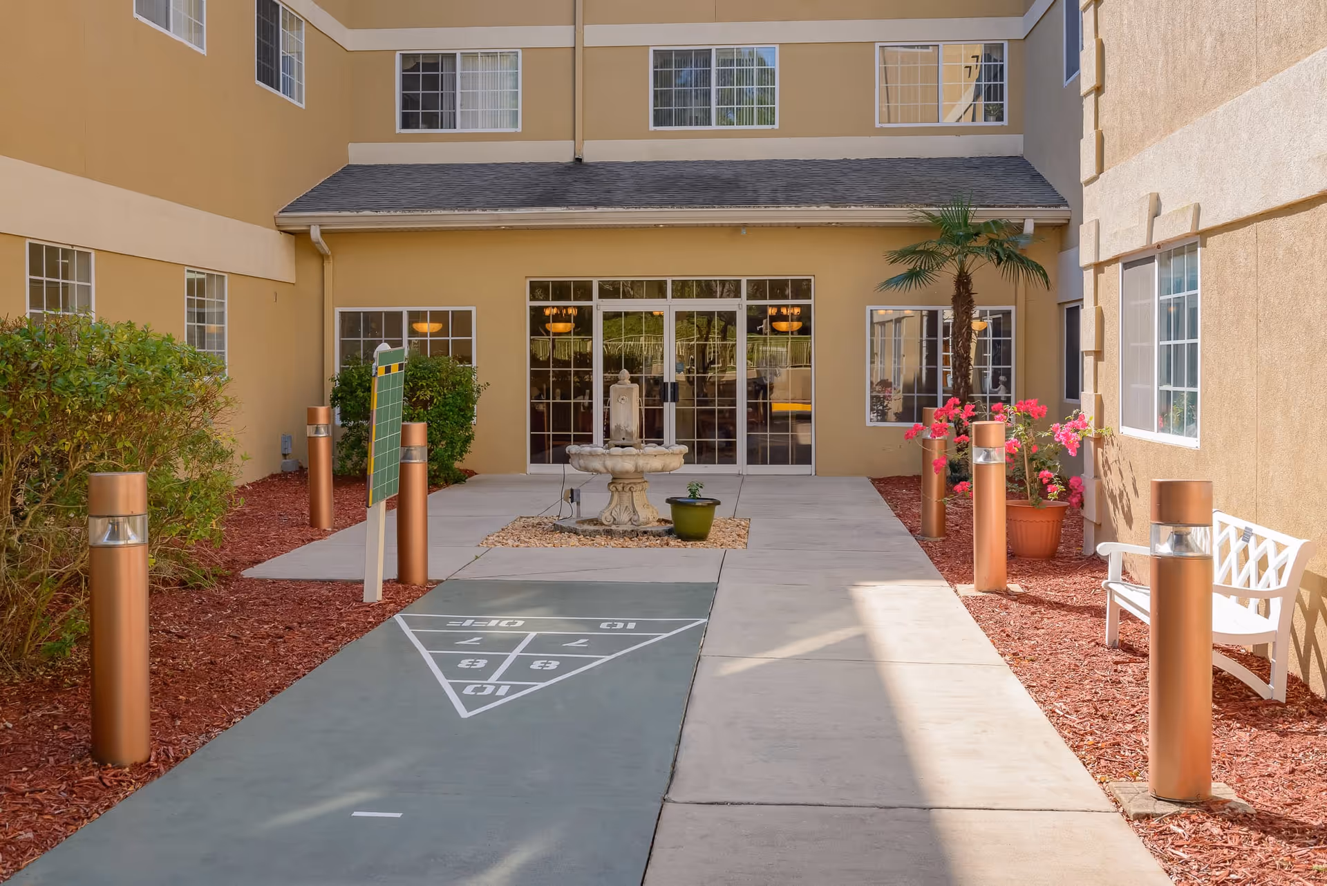 Outdoor courtyard with a shuffleboard court leading to a central fountain and glass entrance doors, flanked by planters, benches, and pathway lighting.