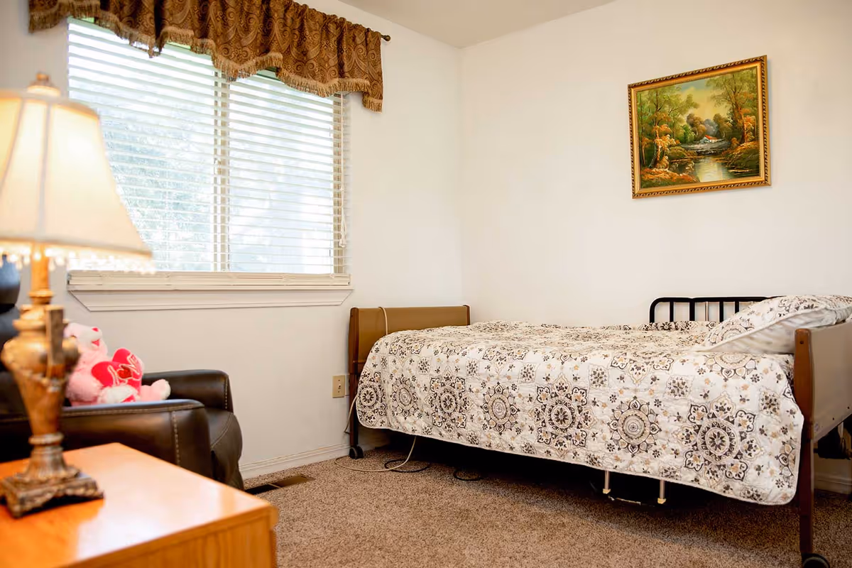 A cozy bedroom with a single bed covered in a patterned quilt, a window with blinds and a brown valance, a black armchair with pink stuffed animals on it, a wooden nightstand with a lamp, and a framed landscape painting on the wall.