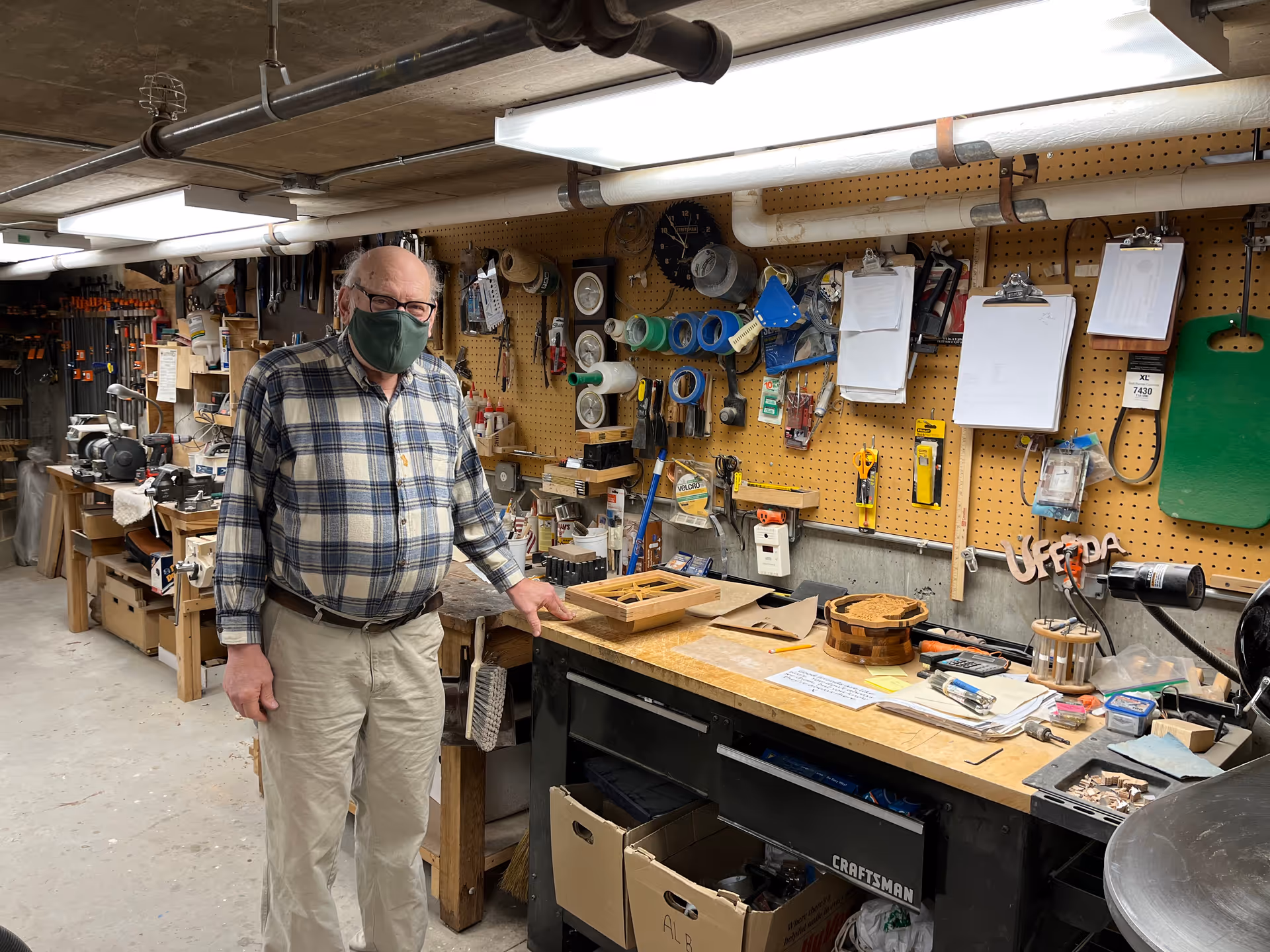 An elderly man wearing a green face mask and a plaid shirt stands in a well-organized workshop with various tools, tapes, and clipboards hanging on a pegboard wall. The workbench in front of him has woodworking projects and supplies, with boxes stored underneath.