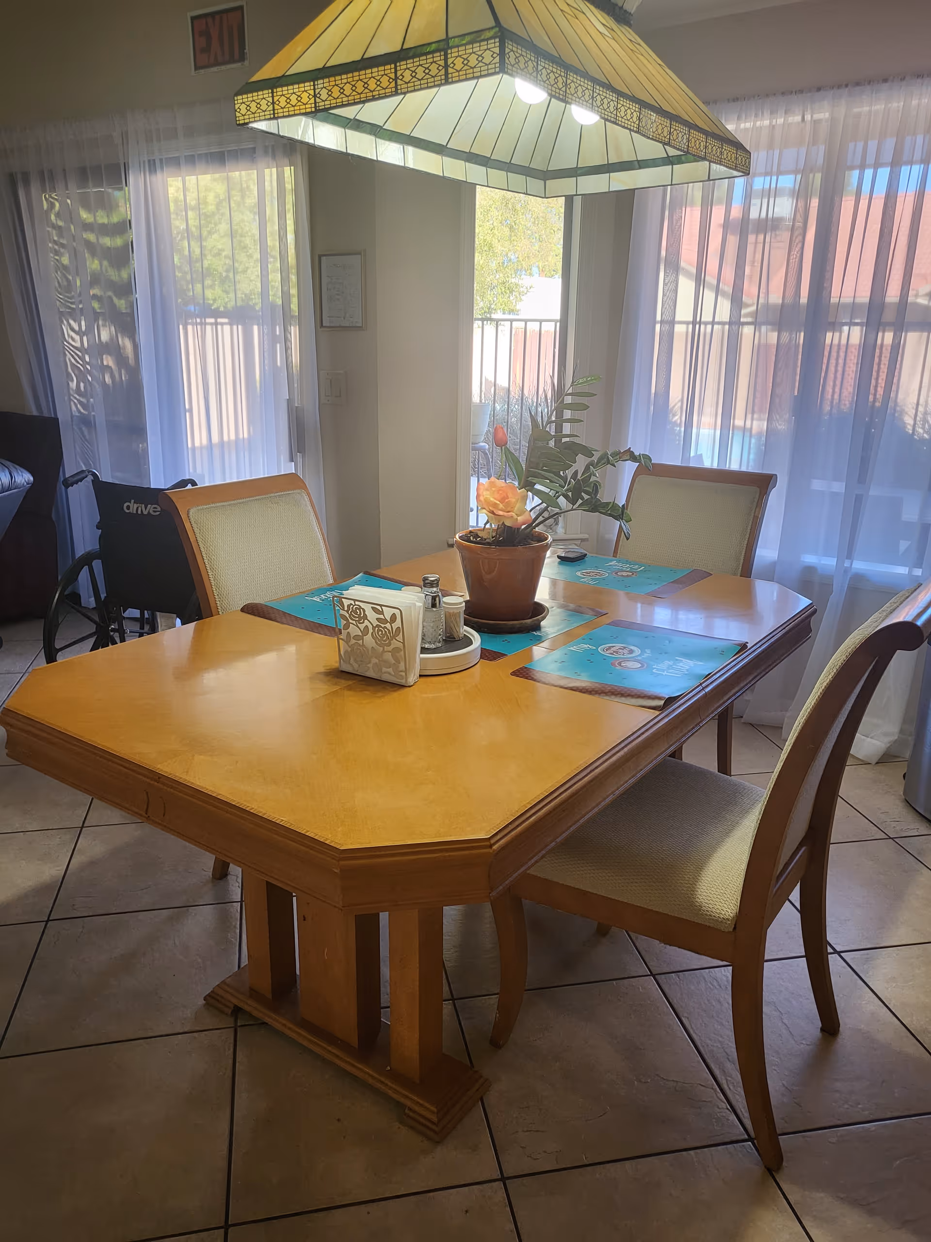 A wooden dining table with four chairs around it, set with blue placemats, a potted plant with a flower centerpiece, and a napkin holder with salt and pepper shakers. A large stained glass pendant light hangs above the table. In the background, sheer white curtains cover large windows letting in natural light, and a wheelchair is partially visible near the left side.