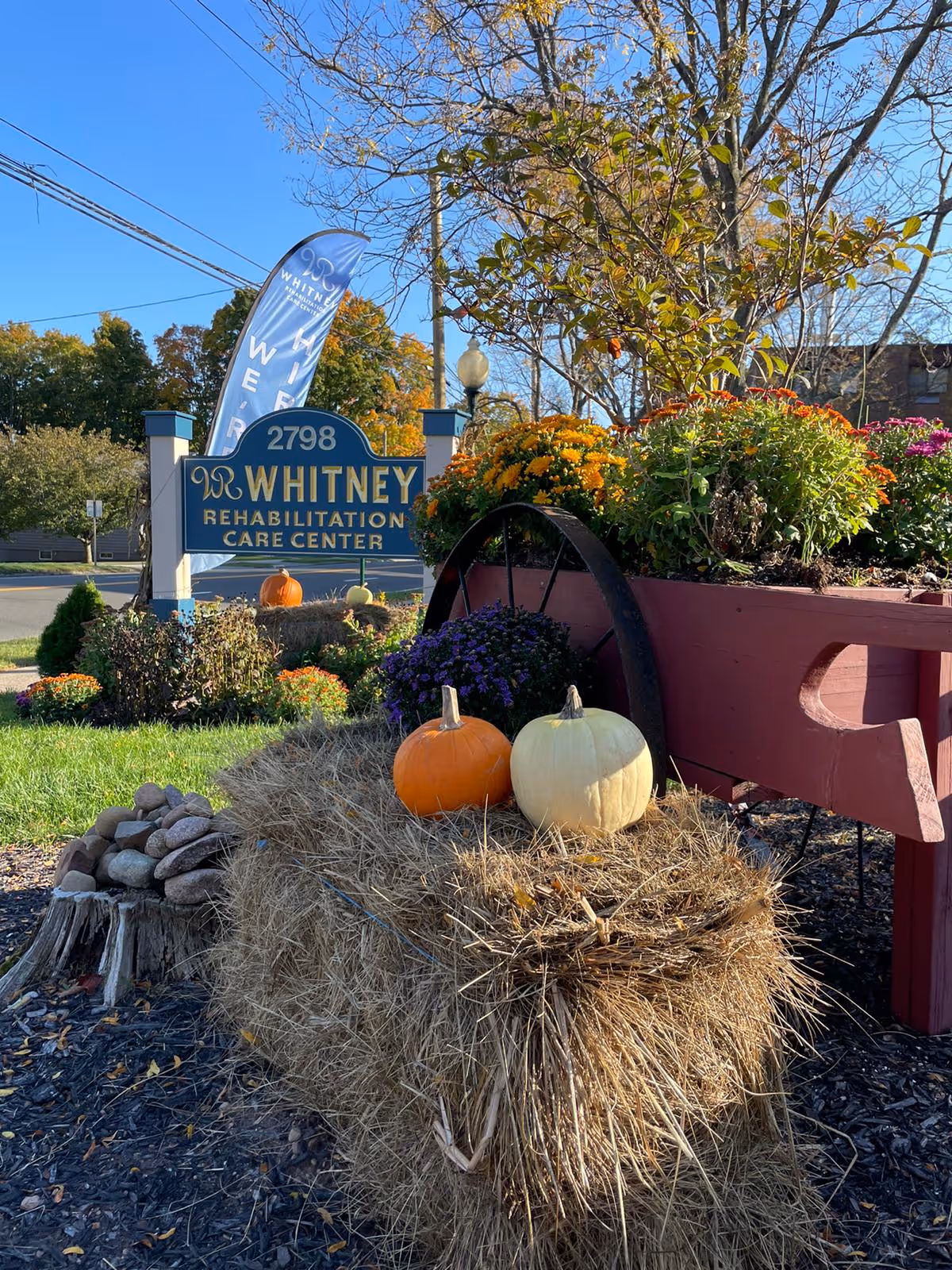 Decorated entrance sign for Whitney Rehabilitation Care Center with pumpkins on a hay bale and a flower-filled wheelbarrow.