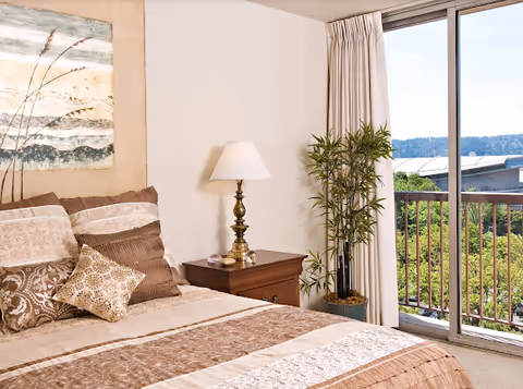 A neatly made bed with multiple decorative pillows in shades of brown and beige, next to a wooden nightstand with a brass lamp and a small plant. A large window with sliding glass doors and white curtains opens to a balcony overlooking greenery and distant hills under a clear sky.