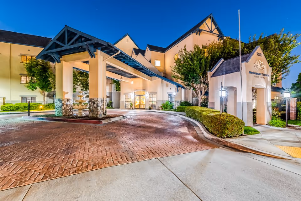 Exterior view of Westminster Terrace Senior Living facility at dusk, showing the entrance with a covered driveway, stone pillars, well-maintained landscaping, and illuminated building facade under a clear blue sky.