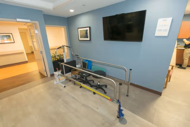 An interior room with blue walls featuring a mounted flat-screen TV and framed artwork. In the center, there is a parallel bar setup used for physical therapy or rehabilitation, with stools placed inside the bars. The room has an open doorway leading to a hallway and another room where a person is partially visible.