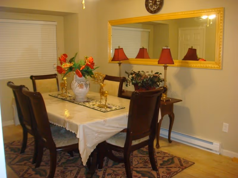 A dining room with a rectangular table covered with a white tablecloth, surrounded by six wooden chairs with cushioned seats. The table is decorated with a vase of red flowers and two gold deer figurines. A large rectangular mirror with a gold frame hangs on the wall above a small side table holding a lamp with a red shade and a potted plant. The room has beige walls, a window with closed blinds, and a patterned rug under the table.