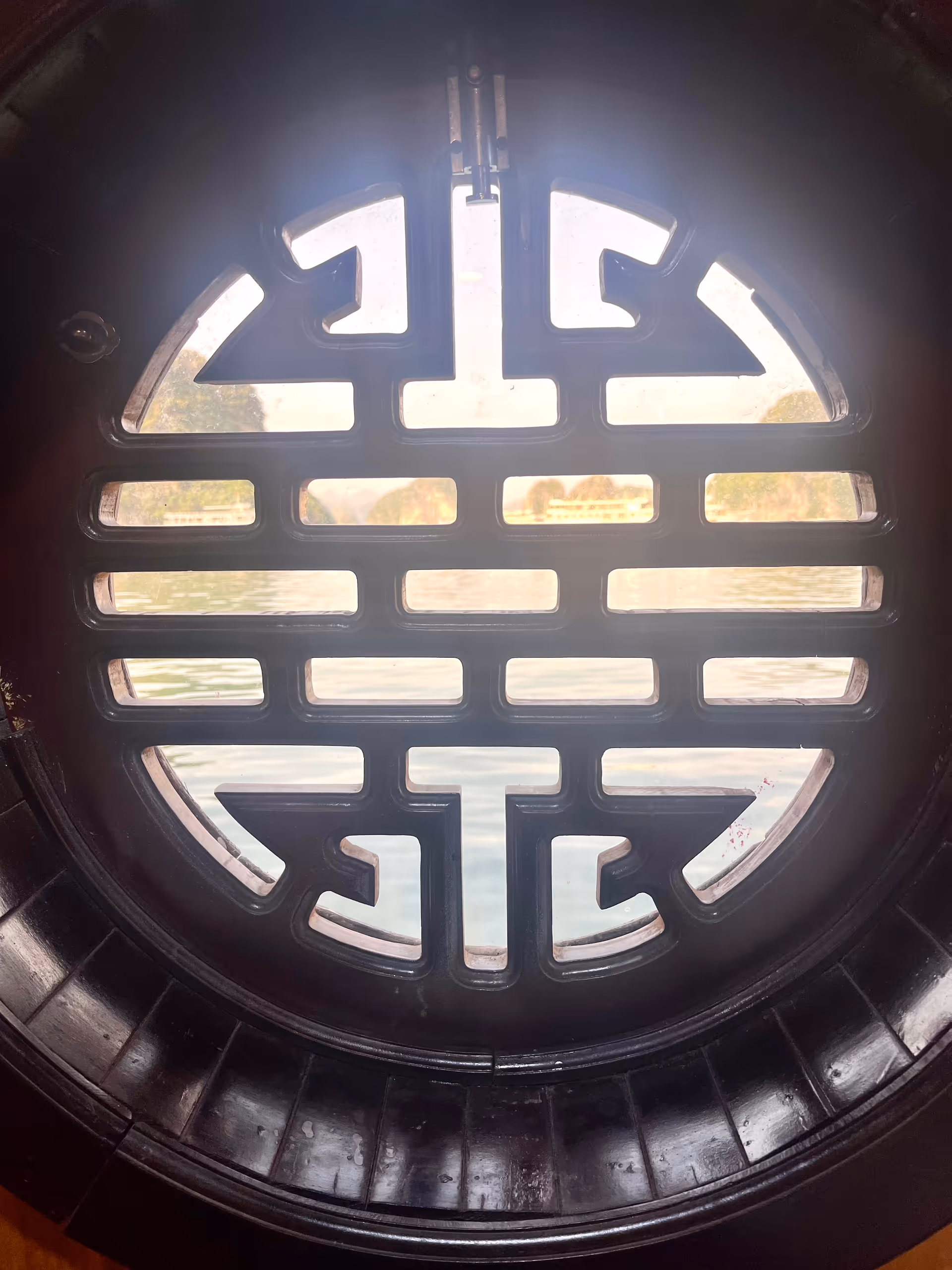 View through a round window with an intricate geometric lattice design, showing a body of water and trees in the background.