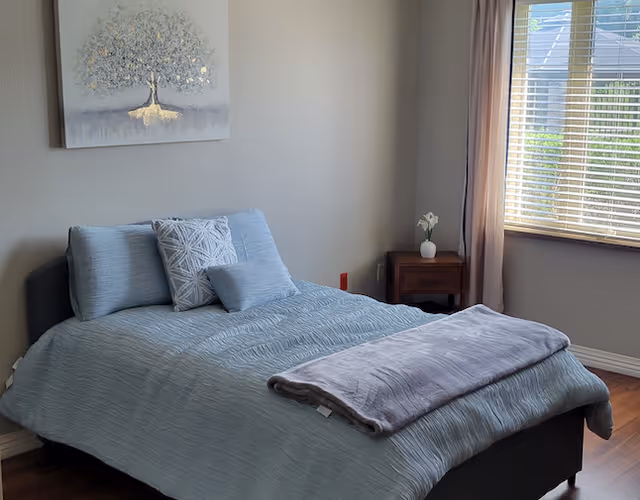 Sunlit bedroom with a neatly made bed dressed in light blue linens, decorative pillows, a nightstand with a small vase, wall art above the bed, and a window with blinds.