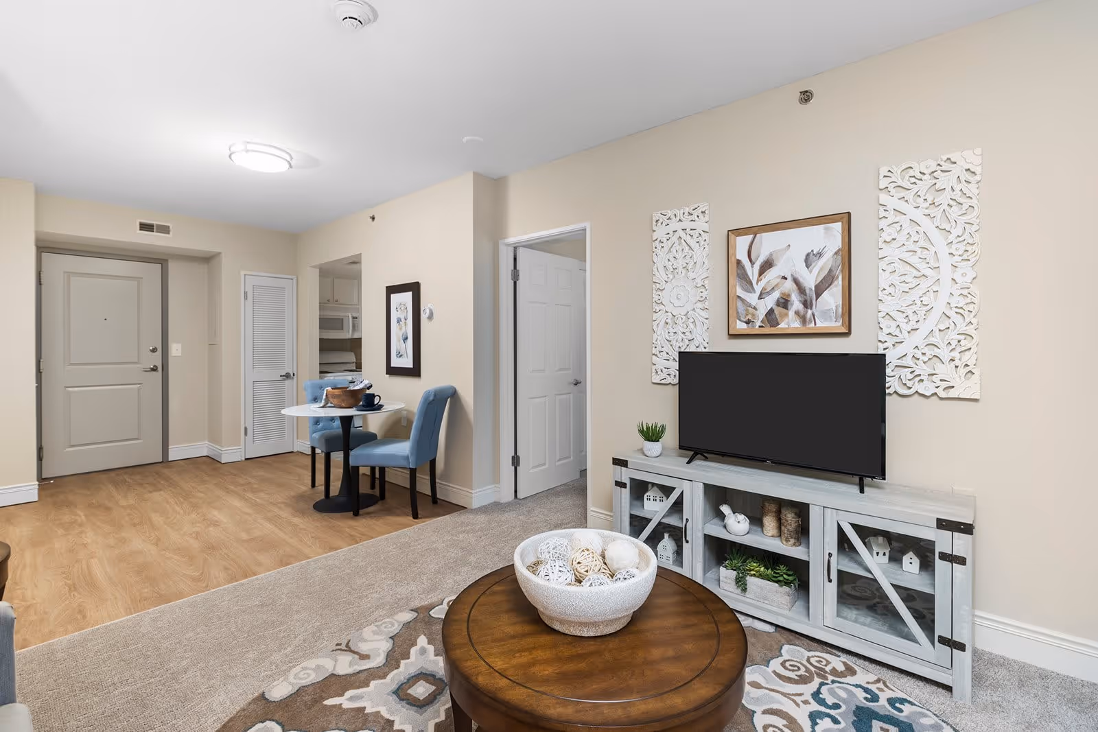 Interior view of a senior apartment living area featuring a round wooden coffee table with decorative balls, a patterned rug, a TV on a rustic white cabinet with plants and decor, two blue upholstered chairs around a small round dining table, light wood flooring, beige walls, and a closed door leading to another room.