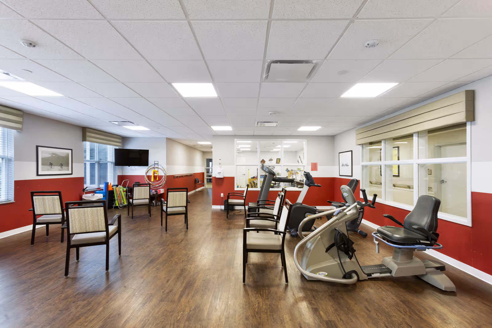 A spacious exercise room with wooden flooring and white ceiling tiles. The room has several chairs arranged in rows and exercise equipment including stationary bikes and treadmills. The walls are painted white with a red lower half, and there are large windows with beige valances. A television is mounted on the wall near the windows, and some exercise accessories are visible in the corner.