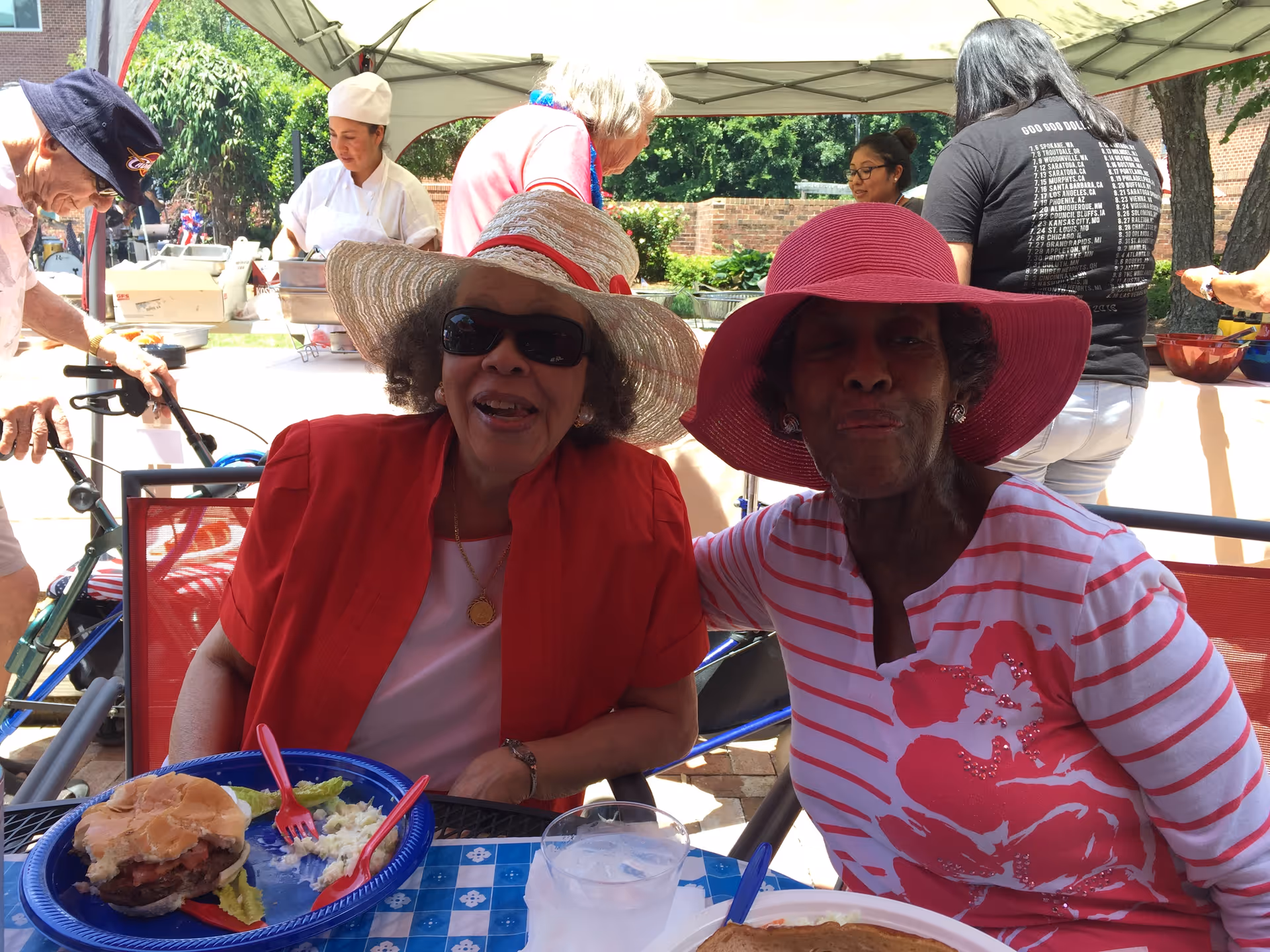 Two elderly women in sun hats sitting at a shaded outdoor table with plates of food during a community event.