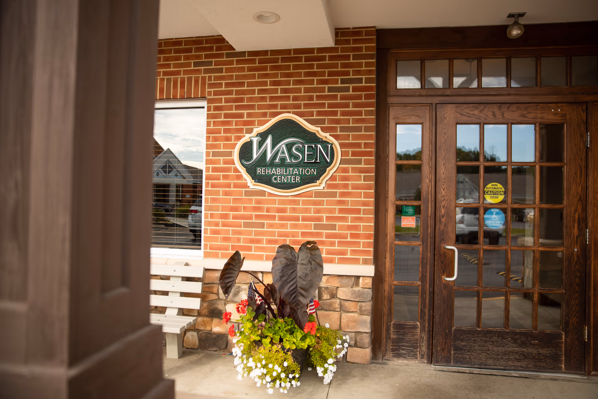 Entrance to Wasen Rehabilitation Center with a brick wall, a wooden door with glass panels, a white bench, and a planter with various plants and flowers.