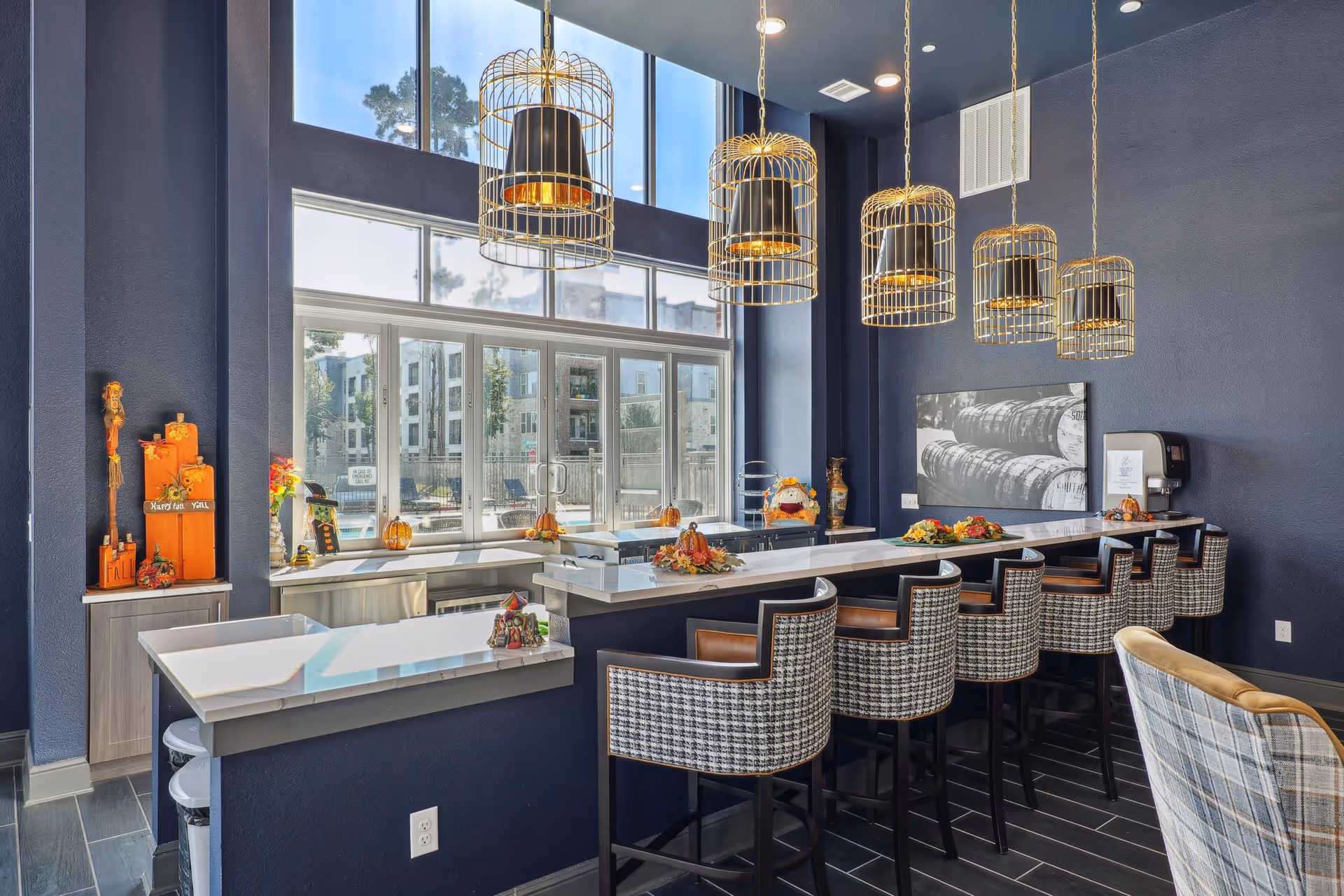 Sunlit communal bar and lounge with a long marble counter, high-backed stools, large windows, and decorative pendant lights.