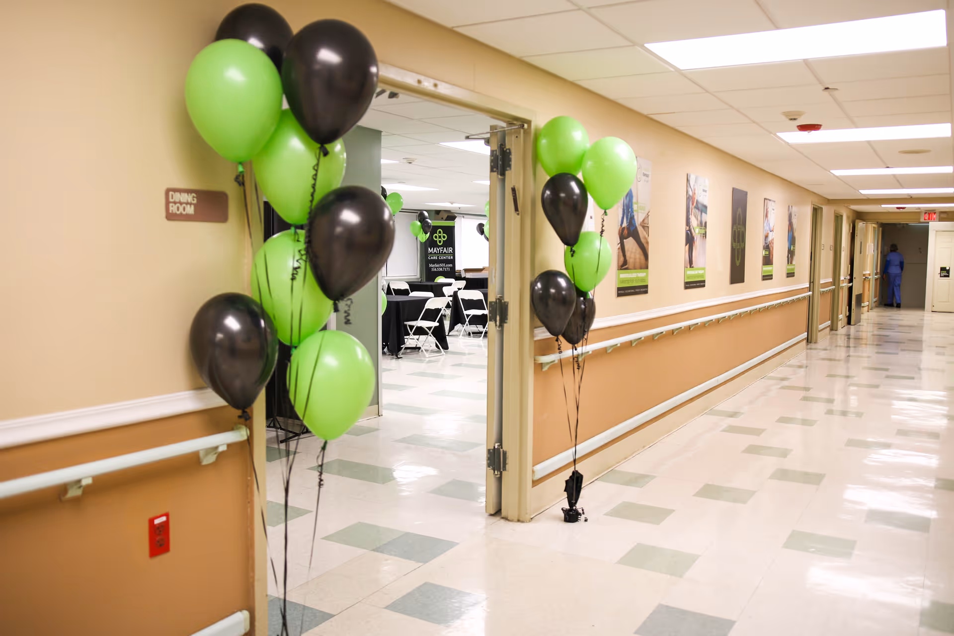 A hallway in Mayfair Care Center decorated with clusters of green and black balloons near the entrance to a dining room. The hallway has beige walls with handrails and several framed posters. Inside the dining room, tables and chairs are arranged, and a banner with the Mayfair Care Center logo is visible.