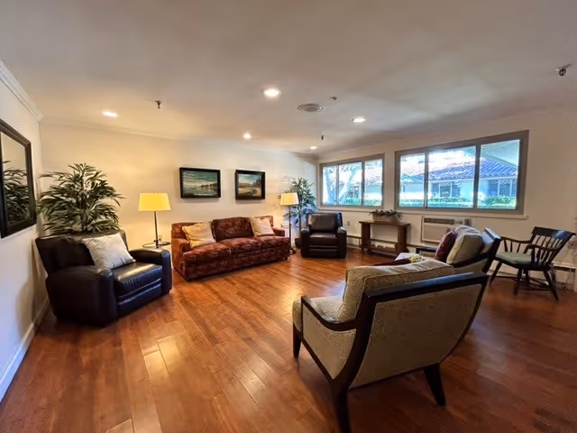 A cozy senior living facility common area with wooden flooring, a patterned sofa, three armchairs, two floor lamps, two framed landscape paintings on the wall, large windows letting in natural light, and some potted plants.