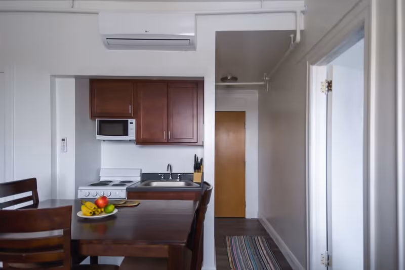 Small apartment kitchenette with wooden cabinets, microwave, sink and stove beside a dining table with a bowl of fruit.