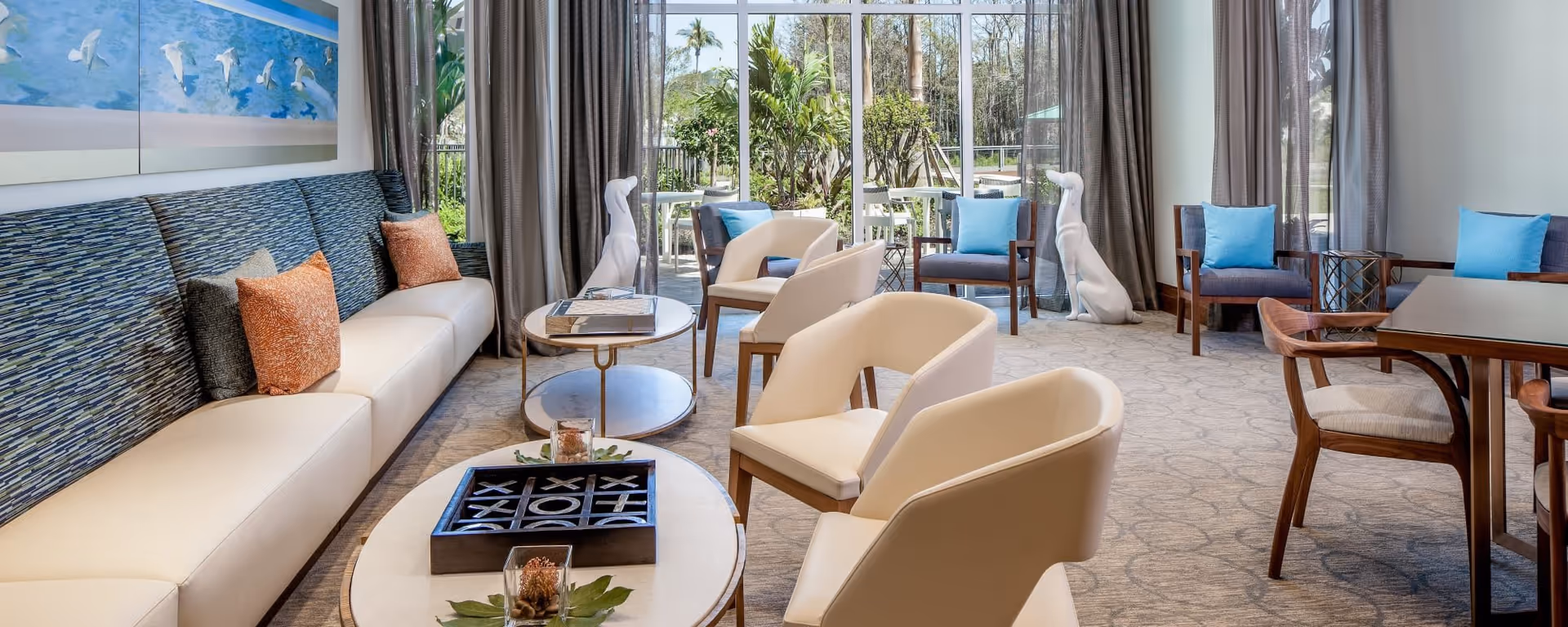 A bright and modern living room area with a long beige sofa adorned with orange and gray pillows, two white armchairs, and several wooden chairs with blue cushions. There are two round coffee tables, one with a tic-tac-toe game and decorative items. Large windows with sheer curtains provide a view of a lush outdoor garden with palm trees. The room features light-colored carpet and wall art depicting birds in flight.