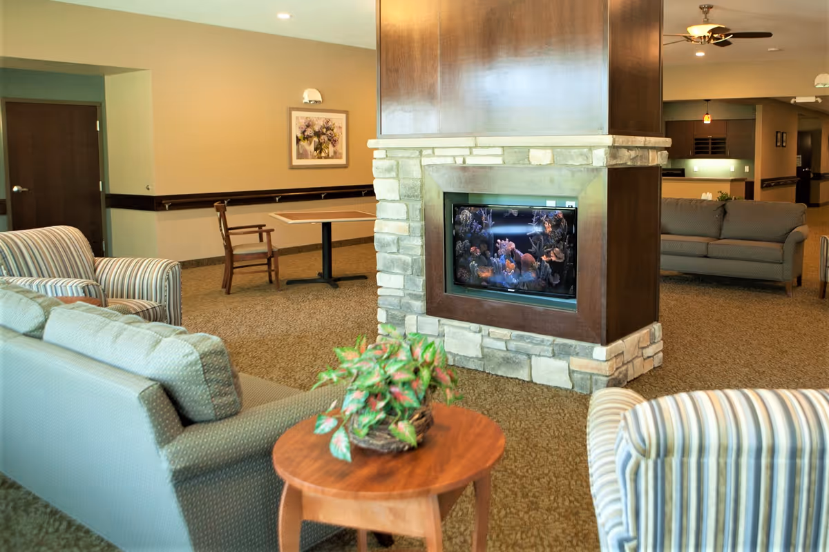 Community living area with upholstered sofas and chairs arranged around a central stone fireplace housing an aquarium and a small table with a potted plant.