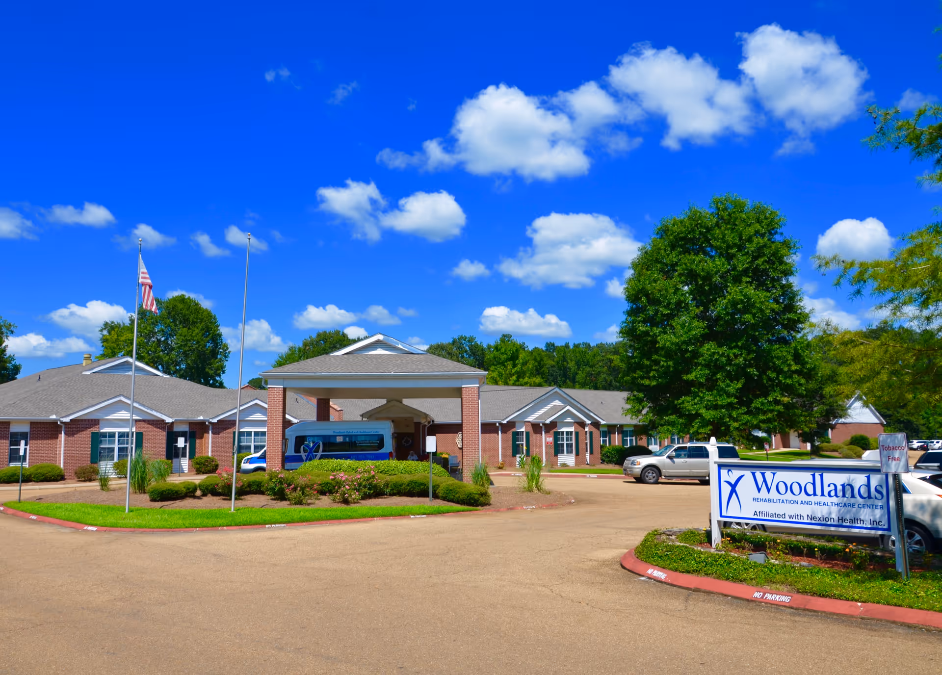Exterior view of Woodlands Rehabilitation and Healthcare Center on a sunny day with a clear blue sky and scattered clouds. The building is a single-story brick structure with a covered entrance where a van is parked. There are trees, shrubs, and a parking area with several cars. A sign in front reads 'Woodlands Rehabilitation and Healthcare Center Affiliated with Nexion Health, Inc.'