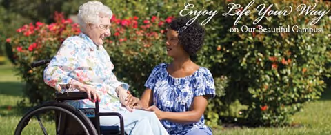An elderly woman in a wheelchair holding hands and smiling with a younger woman kneeling beside her in a garden with green bushes and red flowers in the background.