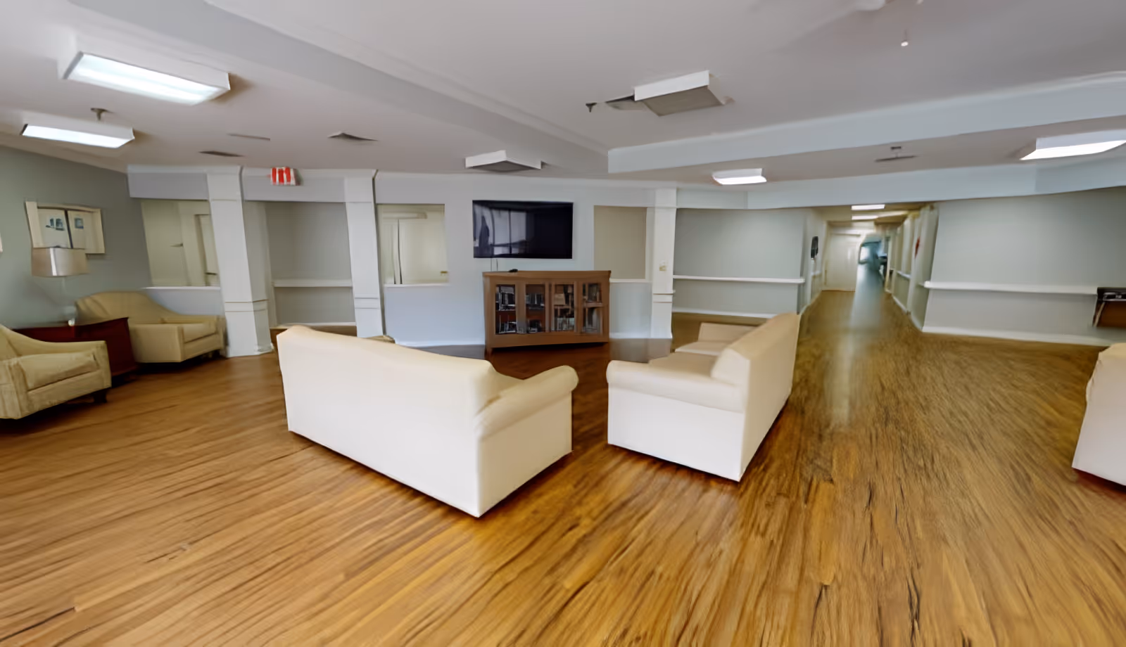 A senior living facility common area with wooden flooring, two cream-colored sofas facing each other, a wooden cabinet with a flat-screen TV mounted above it, and additional armchairs along the walls. The space is well-lit with ceiling lights and has a long hallway extending to the right.