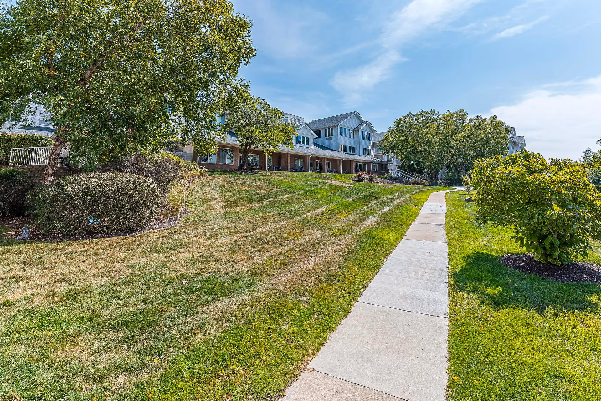 A paved walkway runs through a grassy area with bushes and trees, leading to a large multi-story building with a covered porch. The sky is partly cloudy and the scene is bright and sunny.