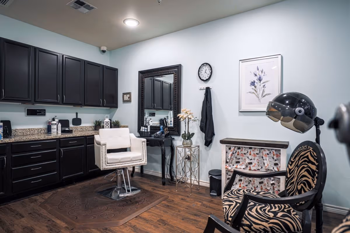 Interior of a salon area with a white salon chair in front of a large mirror, black cabinets with granite countertop, a small table with a vase of flowers, a wall clock, a framed floral picture, and a zebra-patterned chair with a hair dryer hood.