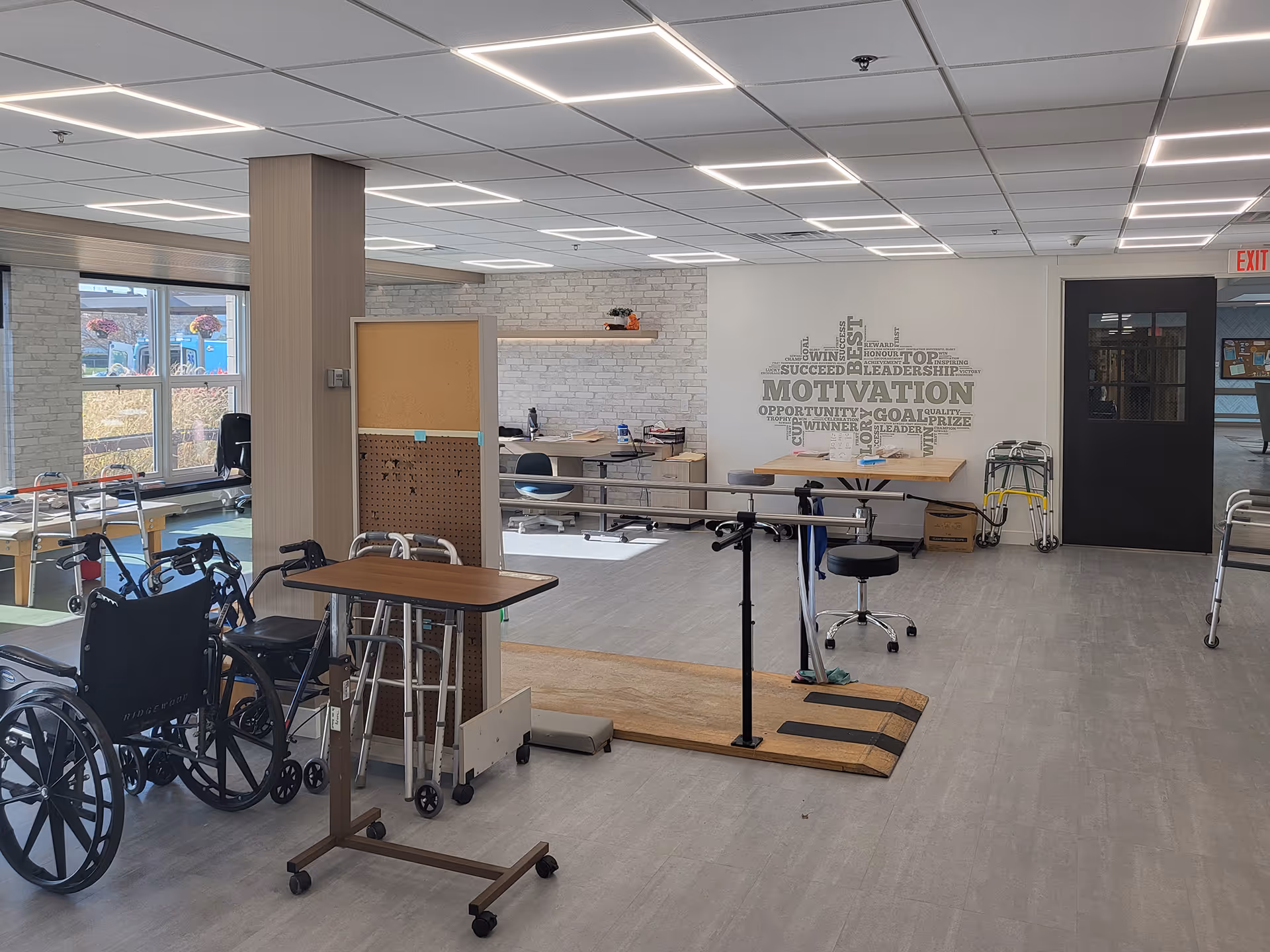 Open rehabilitation/therapy room with parallel bars, wheelchairs, walkers, tables and motivational wall art.