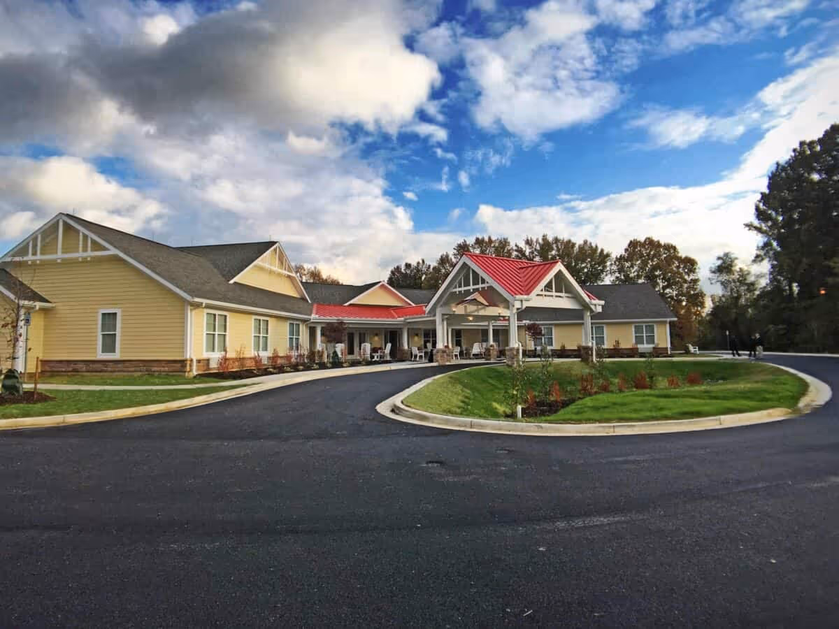 Exterior view of a single-story senior living facility building with yellow siding and a red roof over the entrance. The building is surrounded by a paved driveway and landscaped green areas with trees in the background under a partly cloudy sky.