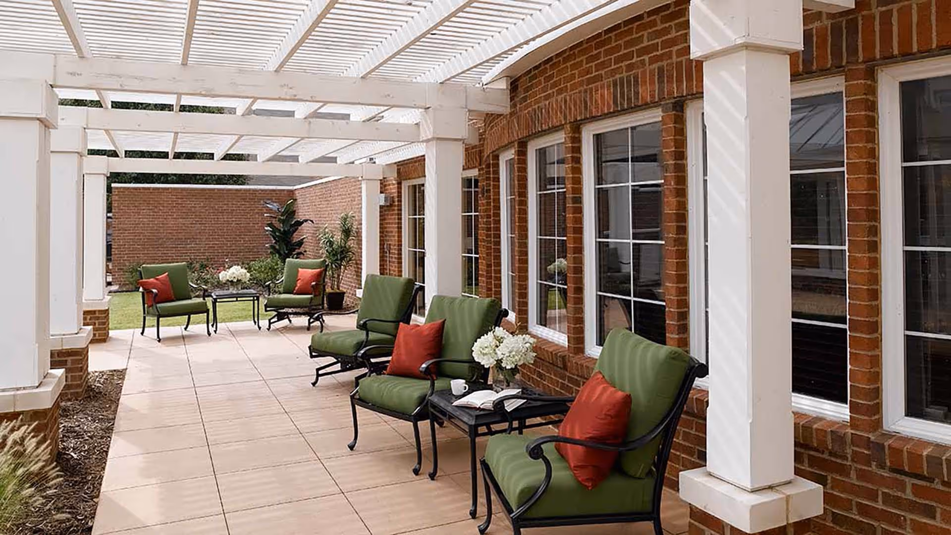 Outdoor patio area with a white pergola overhead, green cushioned chairs with red pillows arranged around small black tables, brick walls with multiple windows, and some potted plants and flowers.