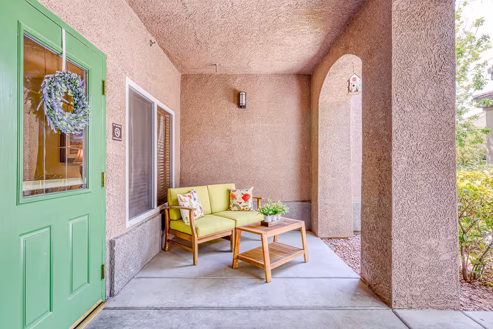 Covered entry porch with a green door, cushioned bench, and wooden coffee table under arched stucco columns.