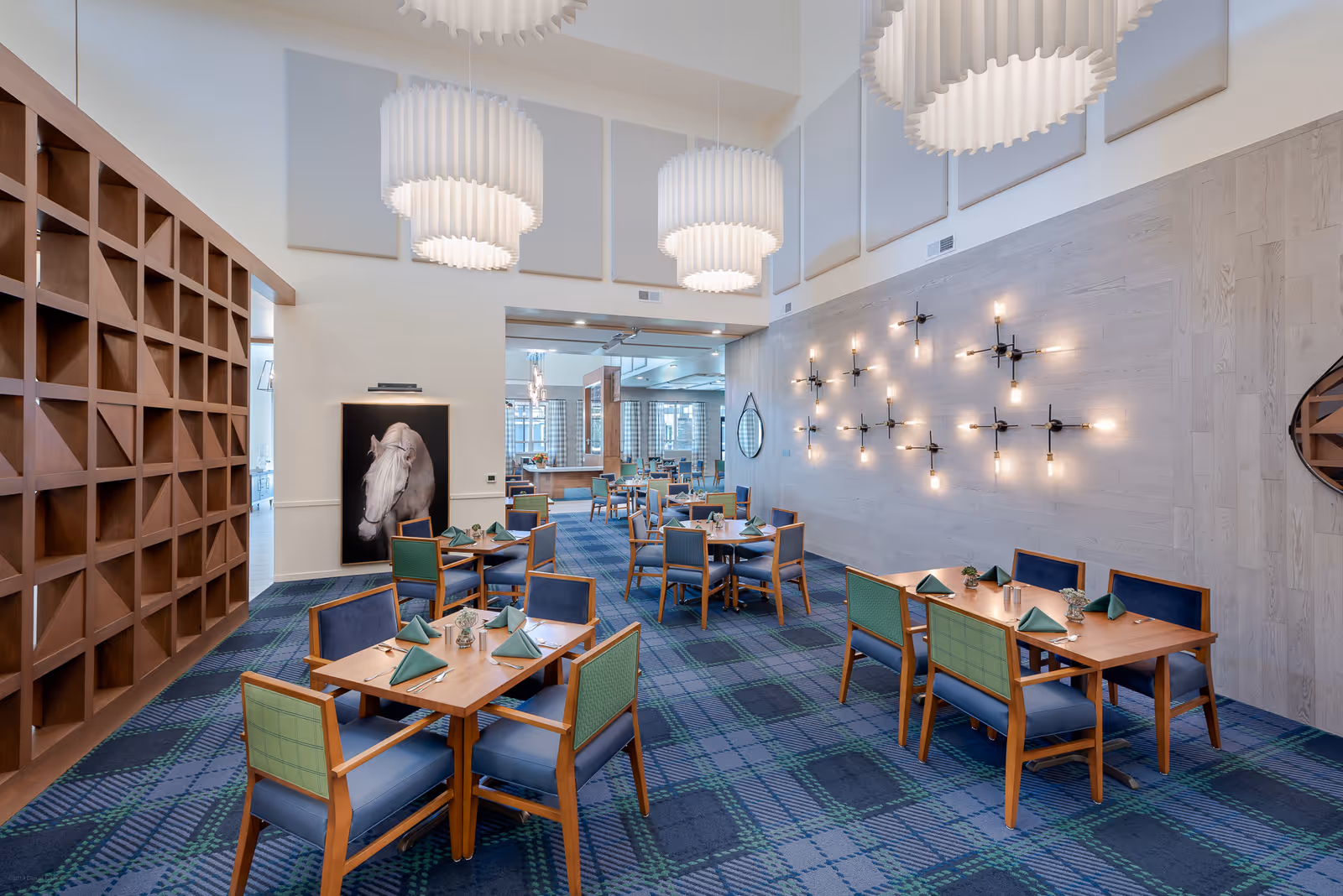 A spacious dining room with wooden tables and chairs, each table set with green napkins and small decorative plants. The room features a blue and green plaid carpet, modern pendant lights hanging from the ceiling, and a wall decorated with multiple light fixtures. A large wooden shelving unit and a framed picture of a white horse are visible on the left side.