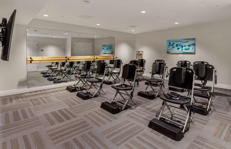 A fitness or exercise room with several black folding chairs equipped with resistance bands and footrests arranged in rows on a patterned carpet. The room has a large wall mirror with a wooden ballet barre, a wall-mounted TV, a water dispenser, and a colorful abstract painting on the wall.