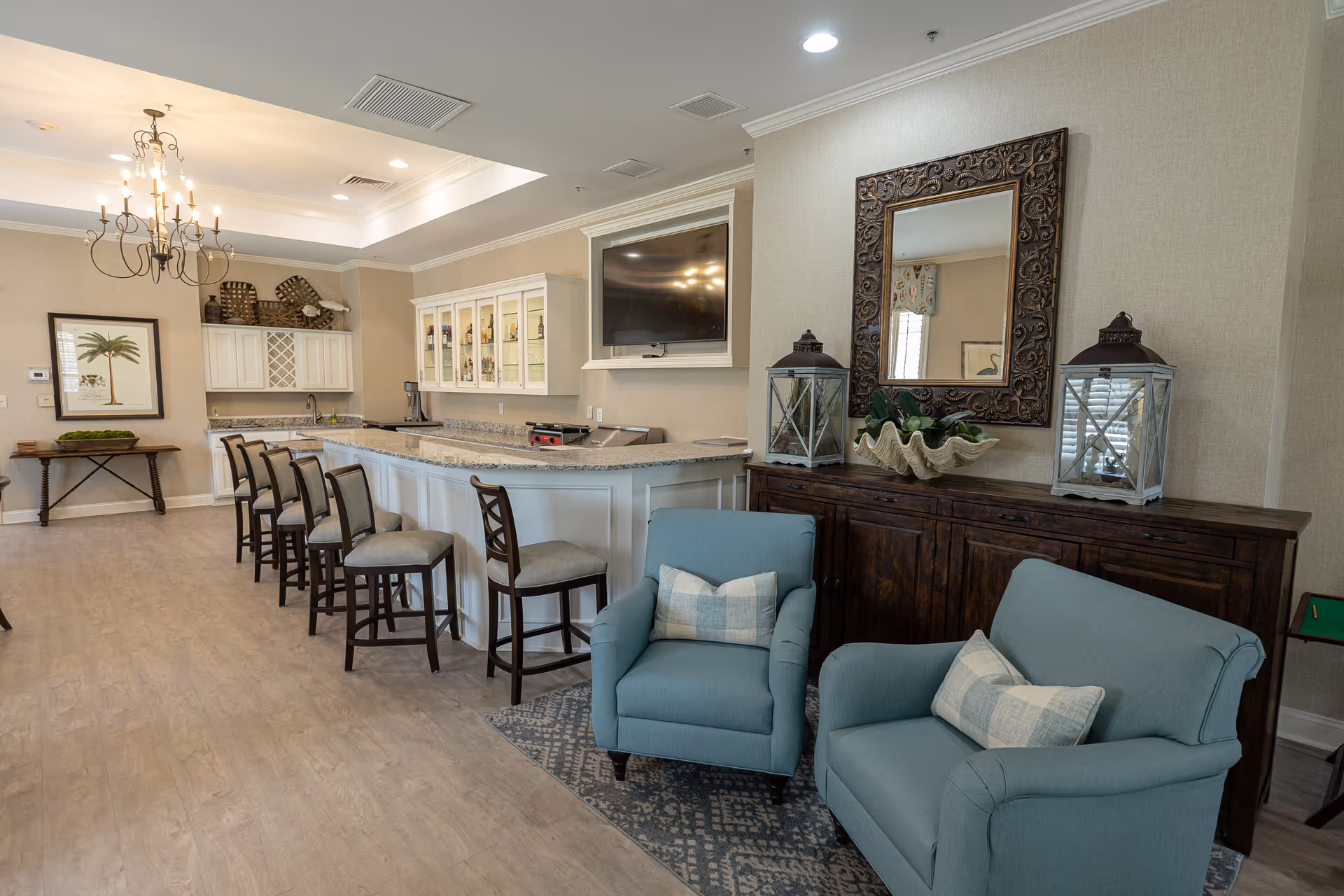 Bright communal lounge and kitchenette featuring a long granite-topped bar with stools, TV, cabinets, and two blue upholstered armchairs.