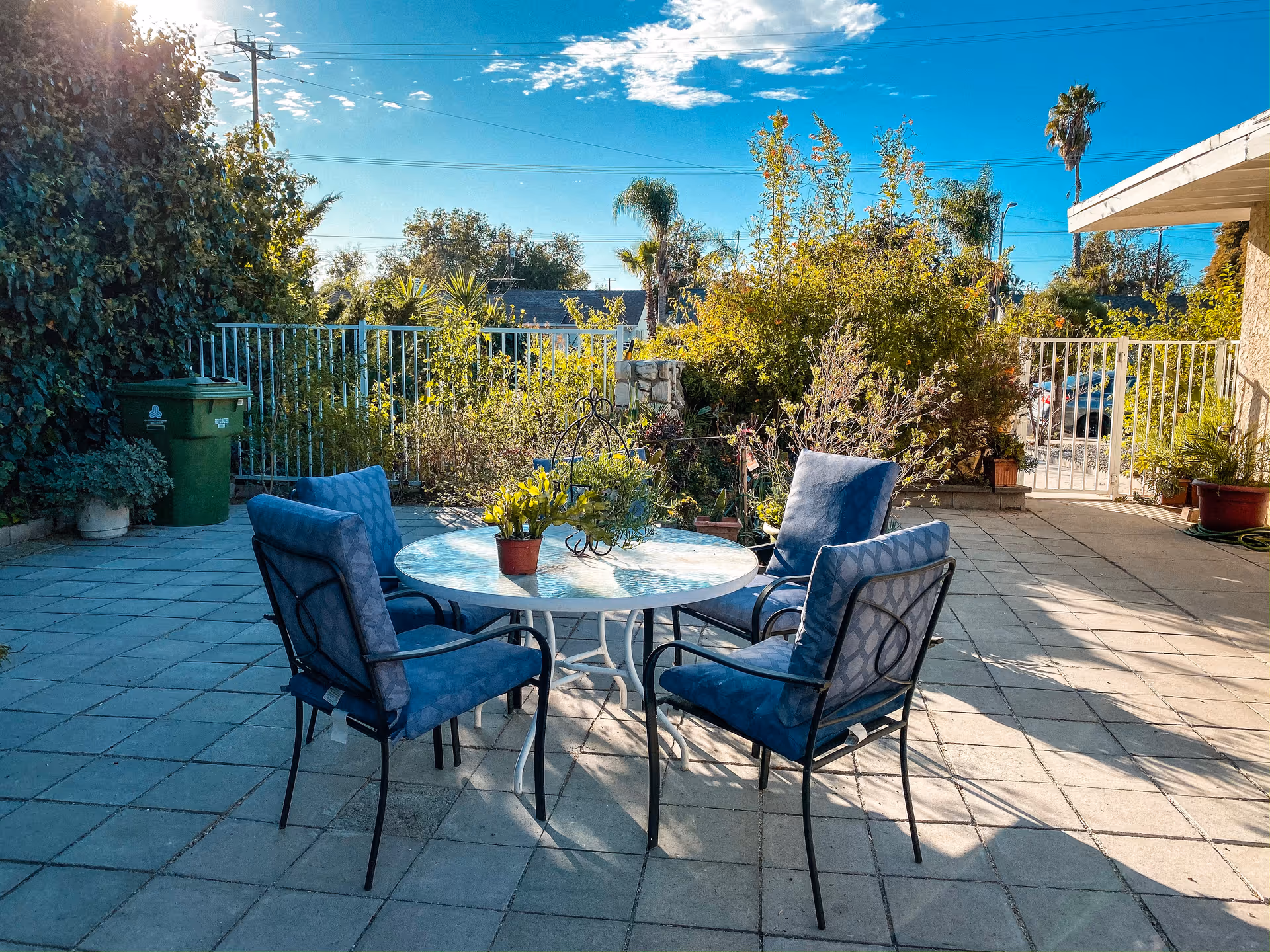 Sunlit outdoor patio with a round table surrounded by four blue cushioned chairs and potted plants.