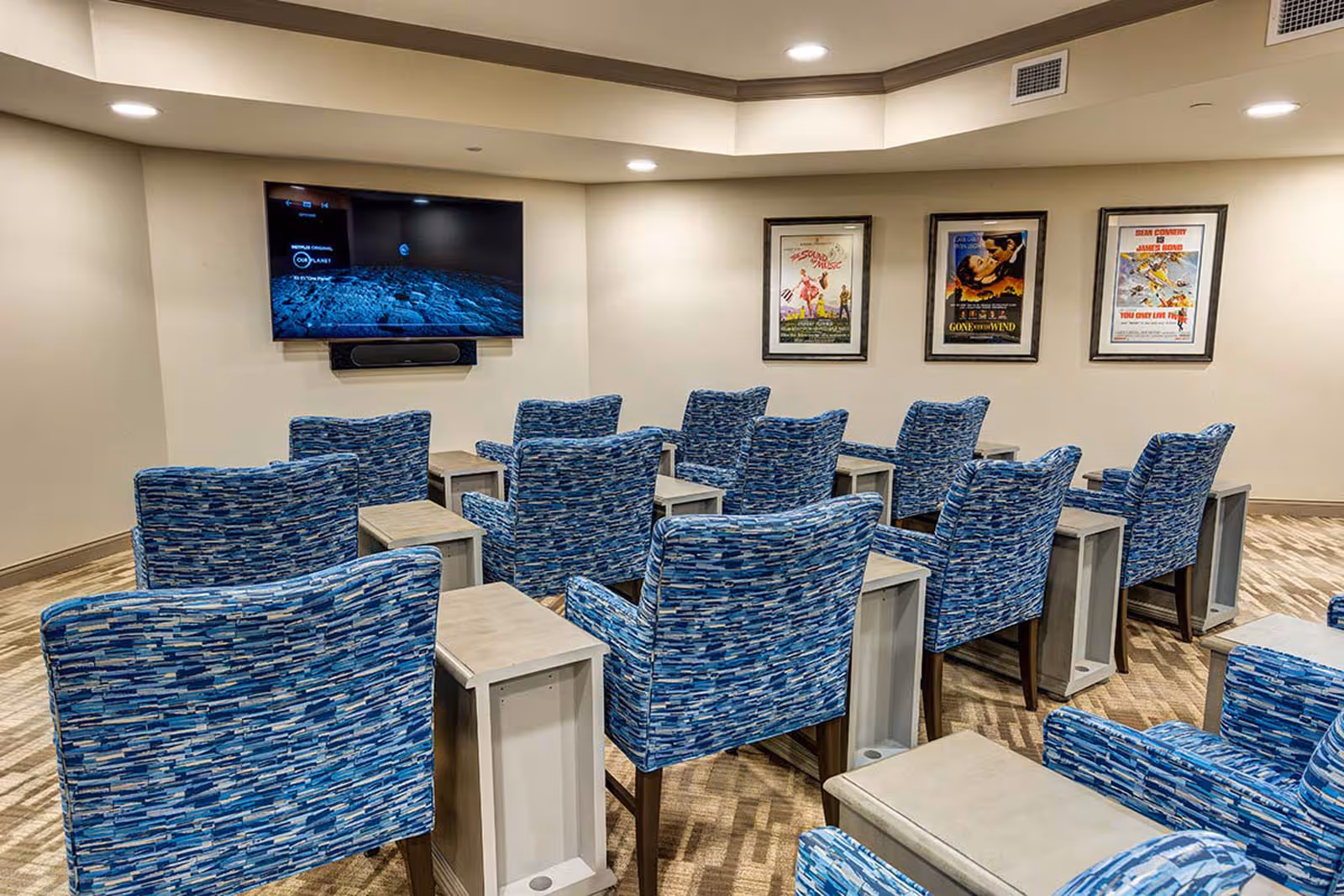 Small community movie room with rows of blue patterned chairs facing a wall-mounted TV and framed posters on the wall.