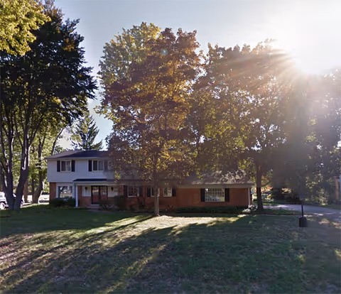 A two-story senior living facility with brick and white siding, set among trees and grass.