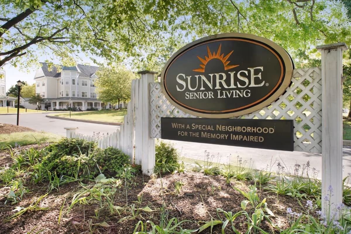 A large Sunrise Senior Living sign by a white lattice fence with landscaped grounds and the facility building visible in the background.