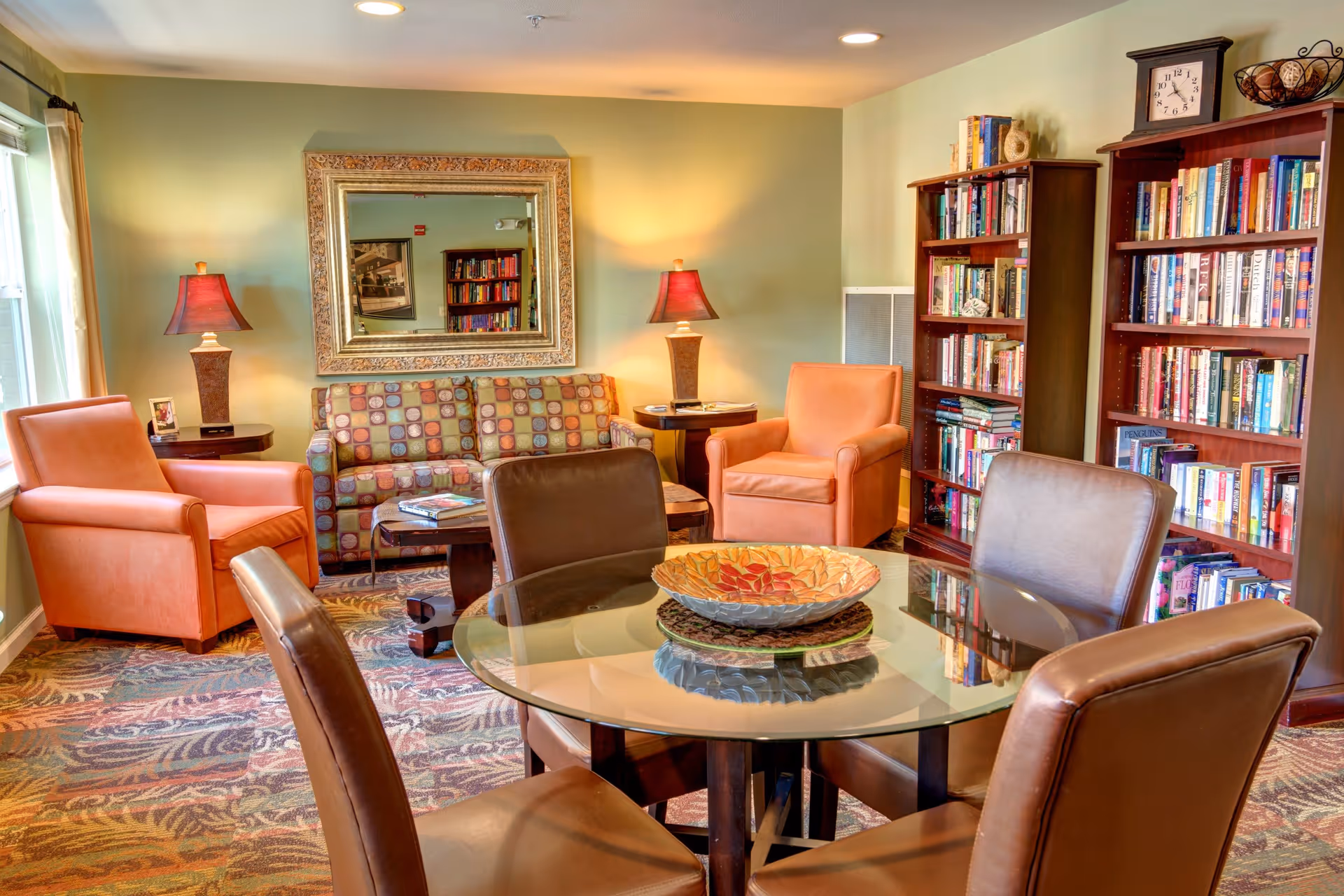 A cozy living room area with a patterned sofa, two orange armchairs, two side tables with lamps, a large decorative mirror on the wall, a glass-top round table with four brown leather chairs, and two tall bookshelves filled with books. The room has a patterned carpet and soft lighting.