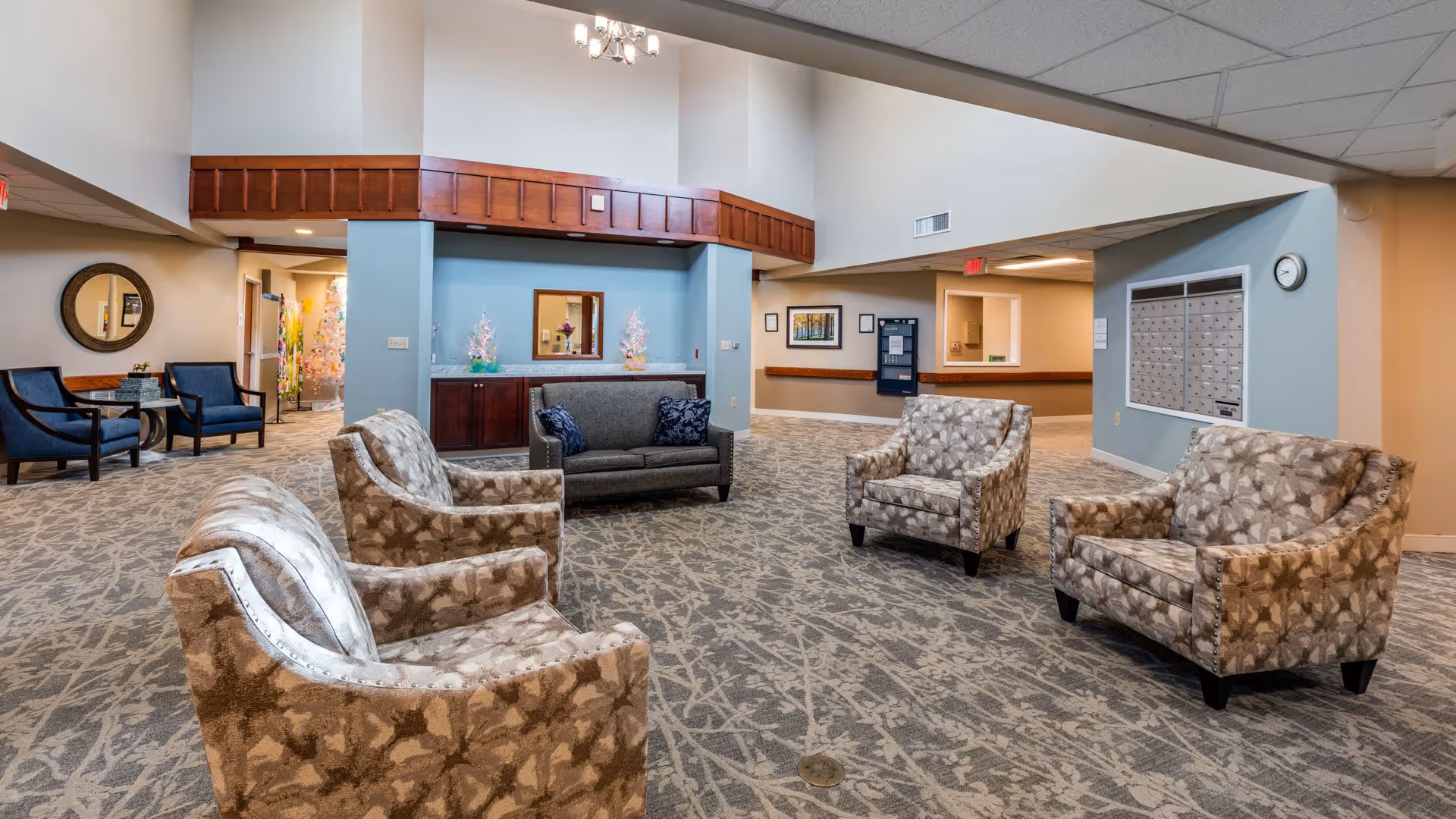 A spacious seating area in a senior living facility with patterned armchairs and a gray sofa arranged on a carpeted floor. The walls are painted light blue and beige, with a wooden trim and a mirror above a cabinet. There are mailboxes mounted on one wall and a clock above them. Additional seating with blue chairs and a round mirror are visible in the background.