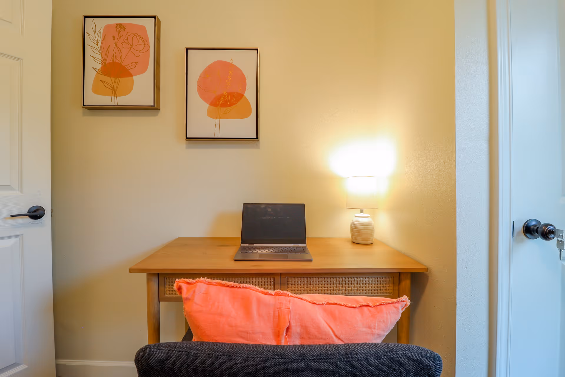 A small wooden desk with a laptop and a lit table lamp on it, positioned against a beige wall. Two framed abstract art pieces with orange and pink shapes hang above the desk. A dark chair with a coral-colored cushion is placed in front of the desk. There are white doors on either side of the desk.