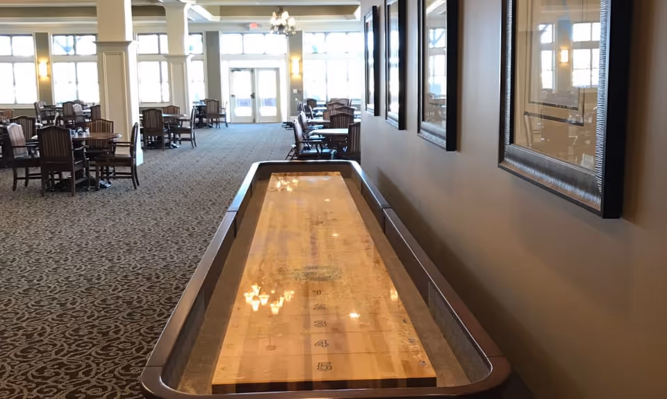 Interior view of a senior living facility common area with a shuffleboard table in the foreground and several wooden tables and chairs arranged in the background near large windows letting in natural light.