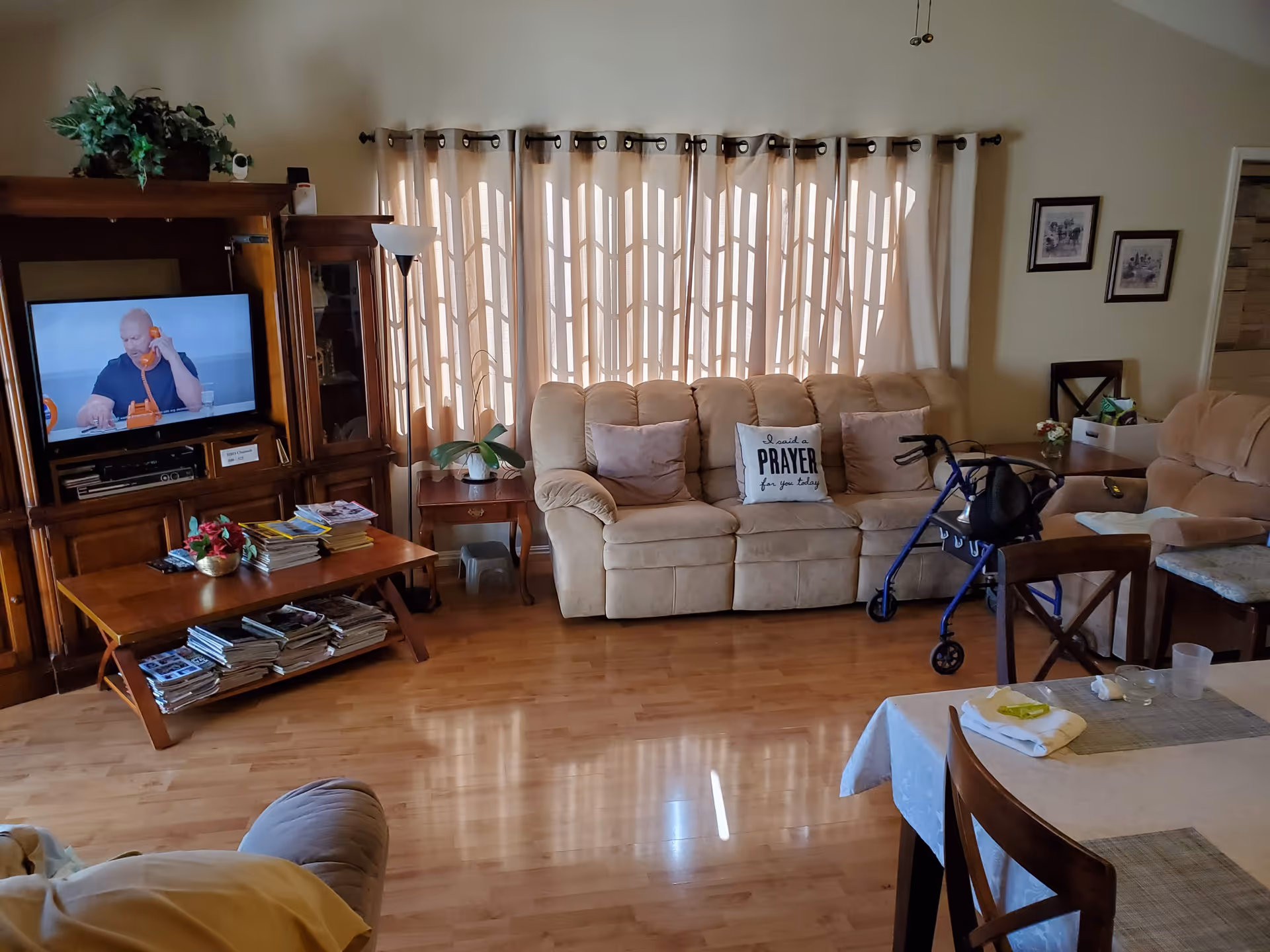 A cozy living room with a beige sofa adorned with pillows, including one that says 'I said a PRAYER for you today.' A blue walker is positioned in front of the sofa. There is a wooden entertainment center with a TV showing a man on the phone, a coffee table with magazines and a small plant, and a dining table with chairs in the foreground. Light filters through beige curtains covering a large window.